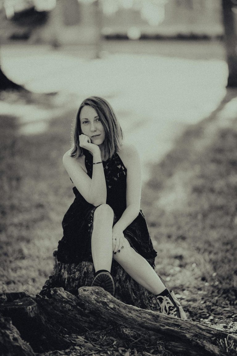 A woman in a black dress sits contemplatively on a tree stump in a tranquil park.