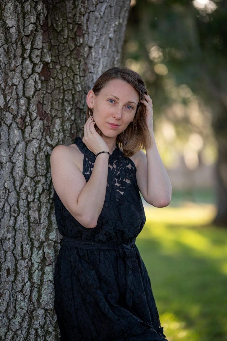 Portrait of a woman in a black dress leaning against a tree in a serene outdoor setting.